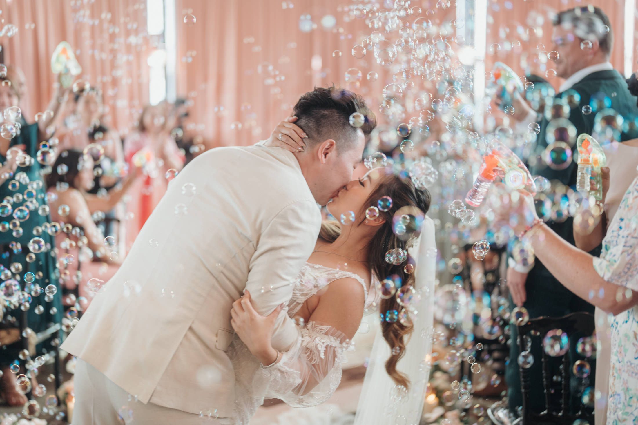 Romantic scene as the newlyweds share a kiss surrounded by bubbles from bubble guns held by joyful guests, creating an enchanting atmosphere of love and celebration