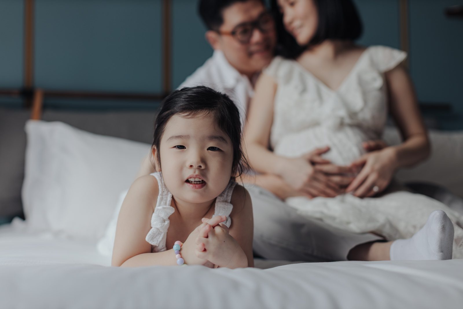 Little girl Naomi in Focus with Parents in Background: Little girl in the foreground lying on the bed, with her parents sitting in the background, smiling