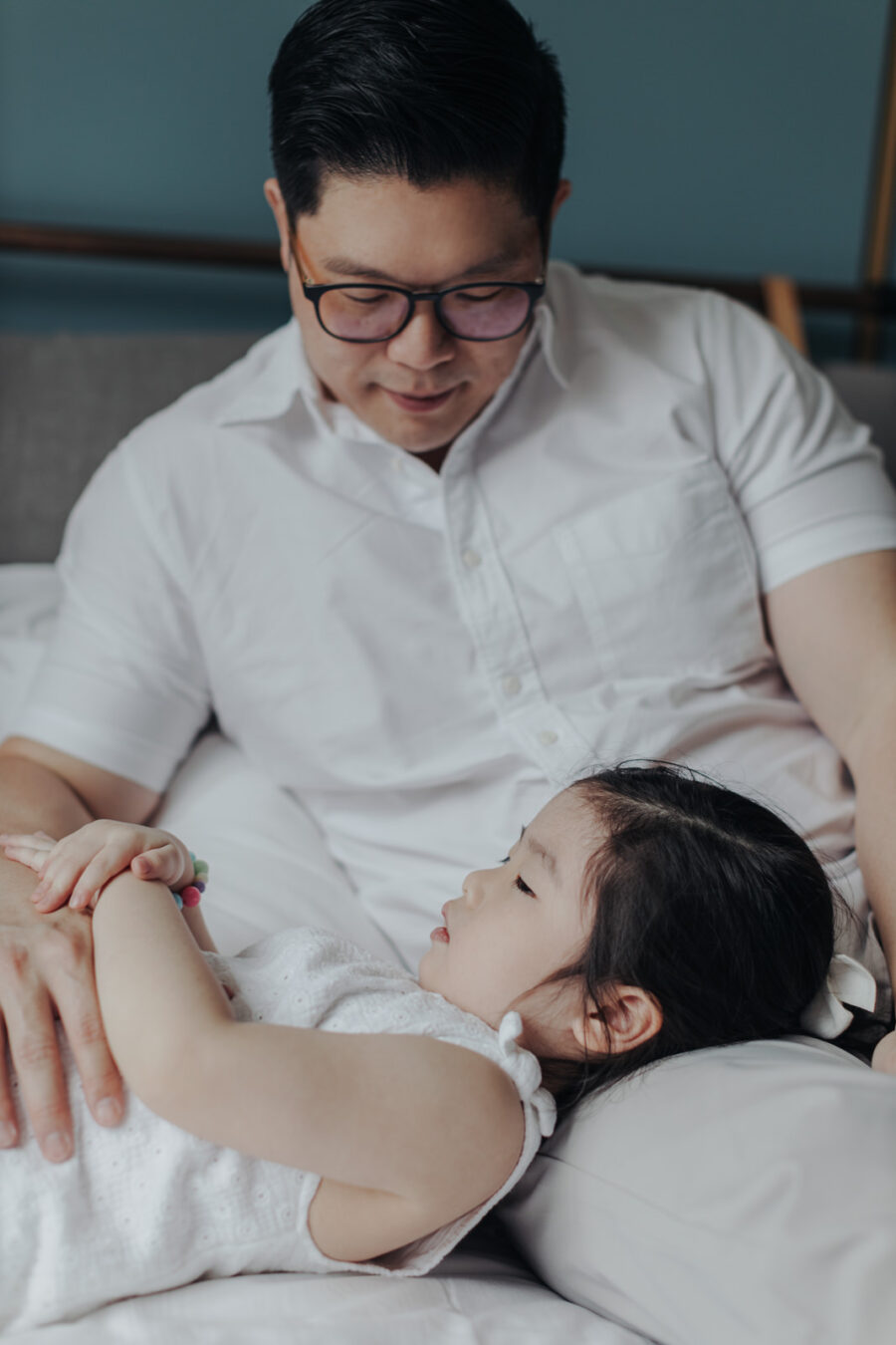 Father and Daughter Bonding: daddy sitting on the bed, lovingly holding and looking at his daughter who is resting on his lap.