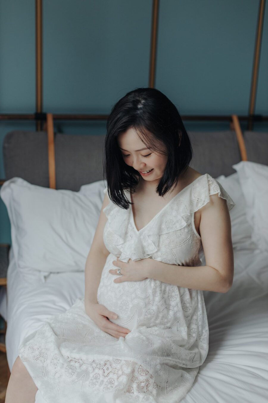 Expecting Mother in White Lace Dress: Mother sitting on the bed in a white lace dress, gently cradling her baby bump and looking at the camera.