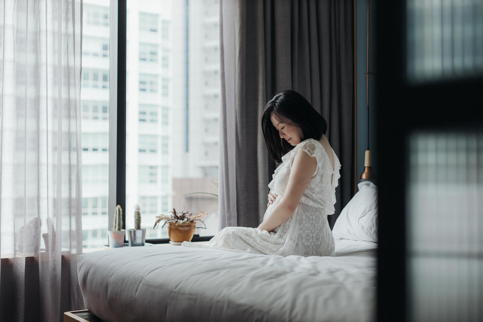 A pregnant wife lying on a bed in a white lace dress, gently cradling her belly. She is looking at the camera with a soft, serene expression. in the room of KLoe Hotel in Kuala Lumpur.