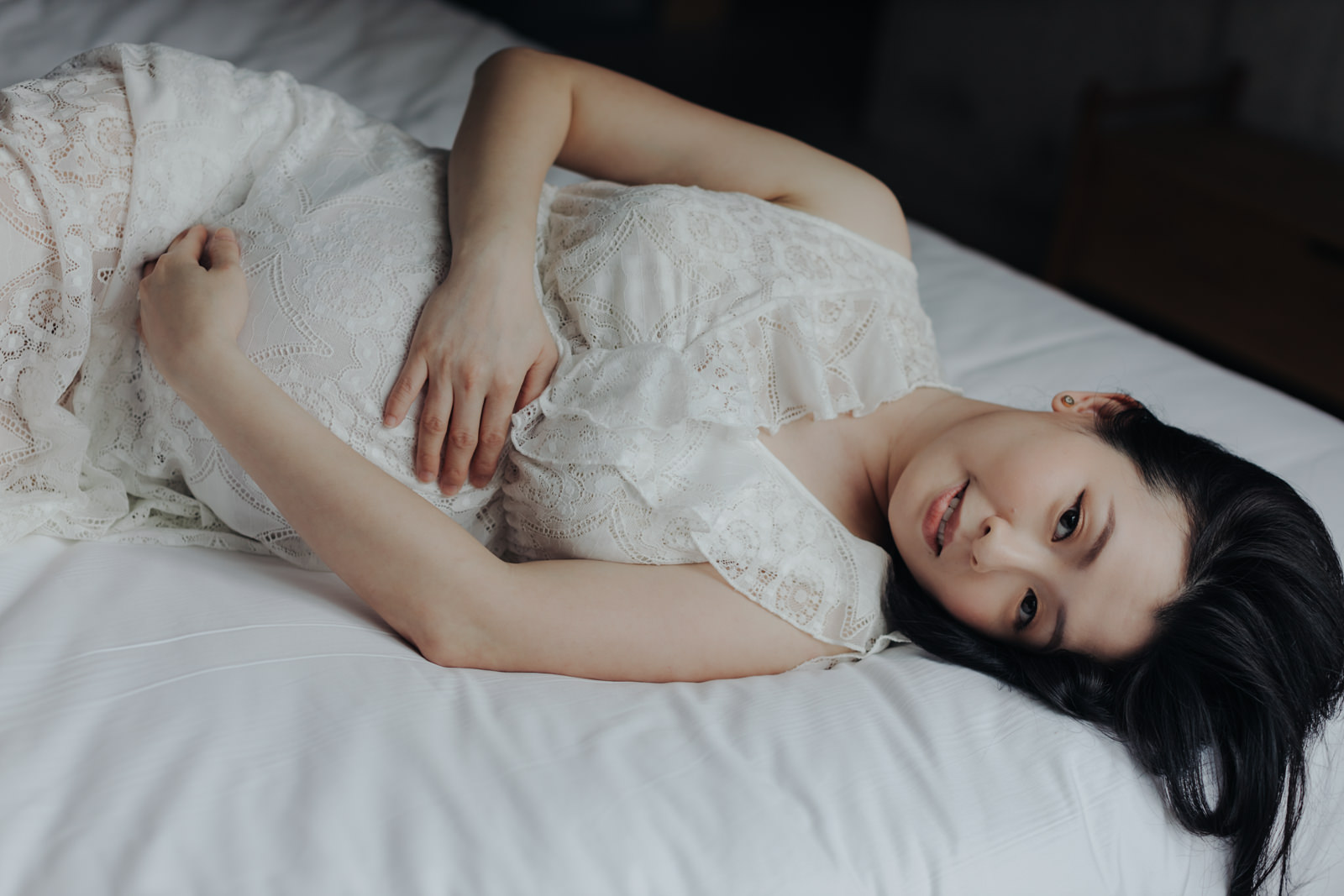 A pregnant wife lying on a bed in a white lace dress, gently cradling her belly. She is looking at the camera with a soft, serene expression during their maternity photo session.