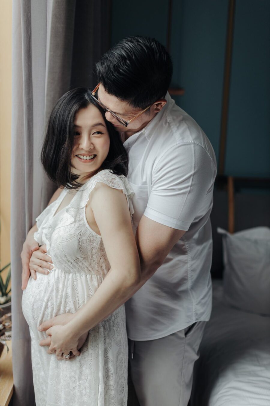 A husband lovingly embracing his pregnant wife from behind. Both are smiling, and she is wearing a white lace dress. The husband gently cradles her belly with his hands.