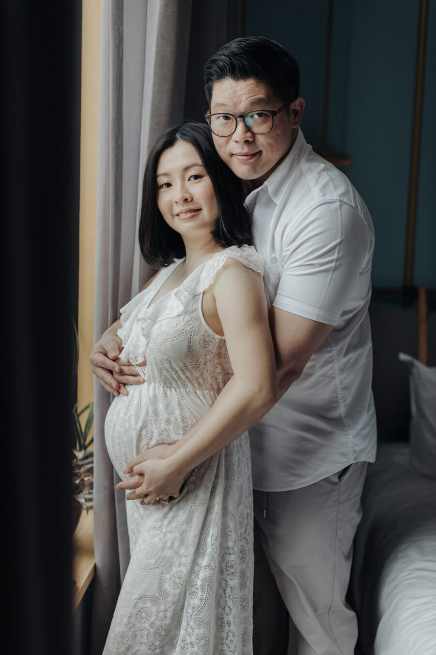 A husband and his pregnant wife standing by the window, looking at the camera. The wife is in a white lace dress, and the husband is standing behind her, both with gentle smiles.