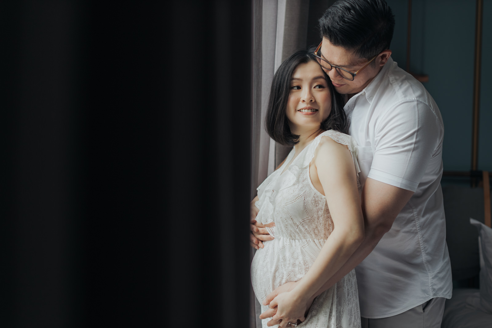 A husband lovingly embracing his pregnant wife from behind. Both are smiling, and she is wearing a white lace dress. The husband gently cradles her belly with his hands in their maternity photo session.