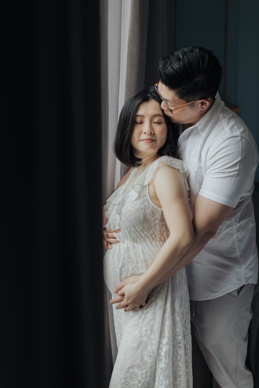 A husband lovingly embracing his pregnant wife from behind. Both are smiling, and she is wearing a white lace dress. The husband gently cradles her belly with his hands highlighting a tender moment in their maternity photo session.