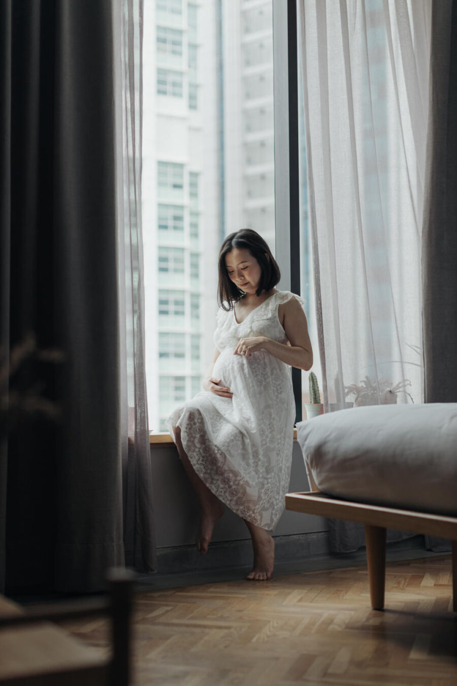 A pregnant wife sitting on a bed by the window in a white lace dress, gently cradling her belly and looking down with a peaceful smile. The cityscape is visible through the window, enhancing their maternity photo session.