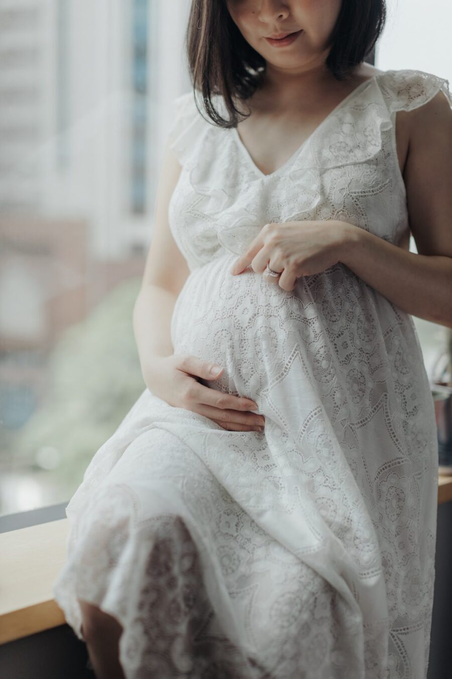 A pregnant wife in a white lace dress sitting by a window, gently cradling her belly. She is looking down with a peaceful smile, and her wedding ring is visible, highlighting a tender moment in their maternity photo session.