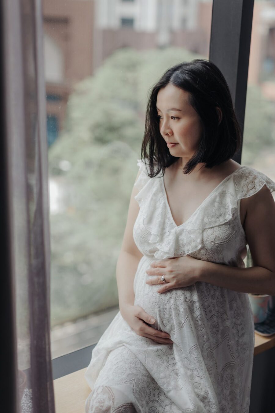 A pregnant wife sitting on a bed by the window in a white lace dress, gently cradling her belly and looking down with a peaceful smile. The cityscape is visible through the window, enhancing their maternity photo session.