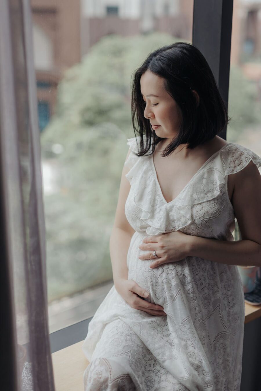 A pregnant wife sitting on a bed by the window in a white lace dress, gently cradling her belly and looking down with a peaceful smile. The cityscape is visible through the window, enhancing their maternity photo session.