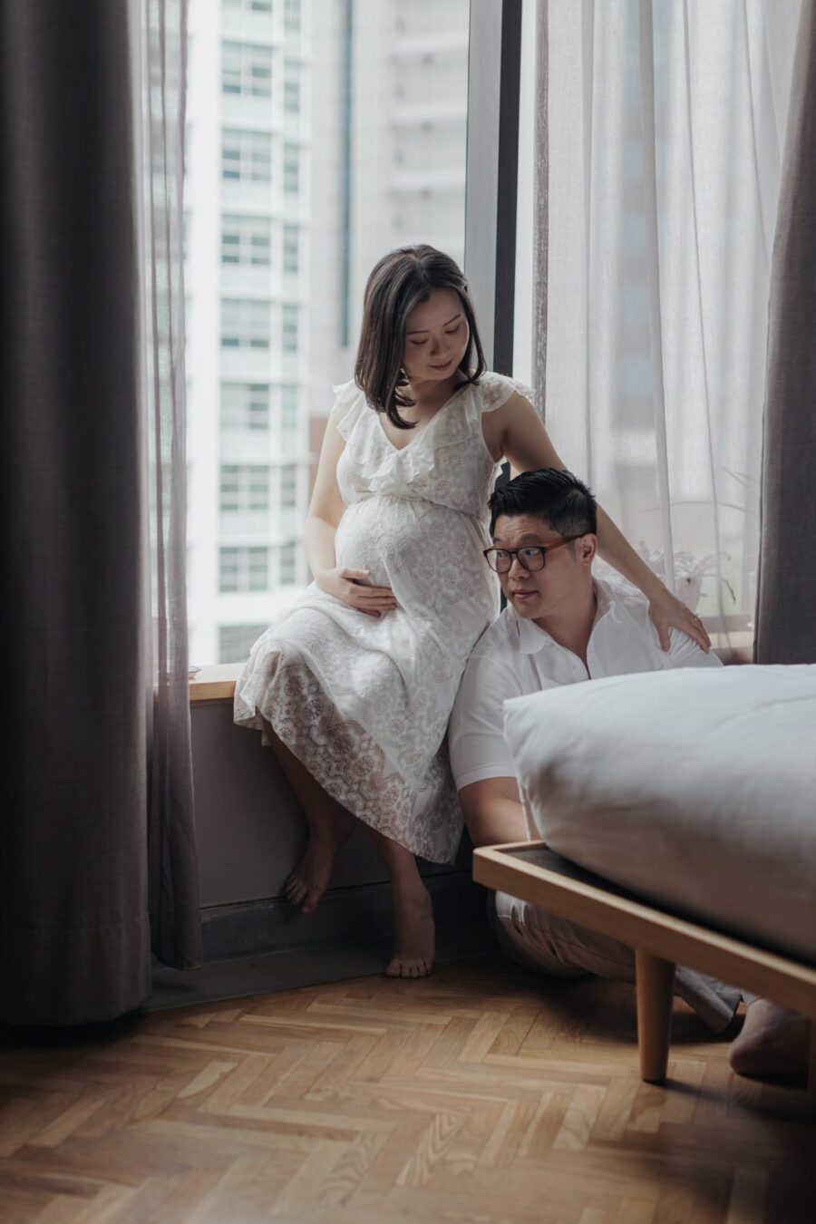 A pregnant wife in a white lace dress sitting on a windowsill, gently cradling her belly, with her husband sitting on the floor beside her. They both have a calm and reflective expression during their maternity photo session.