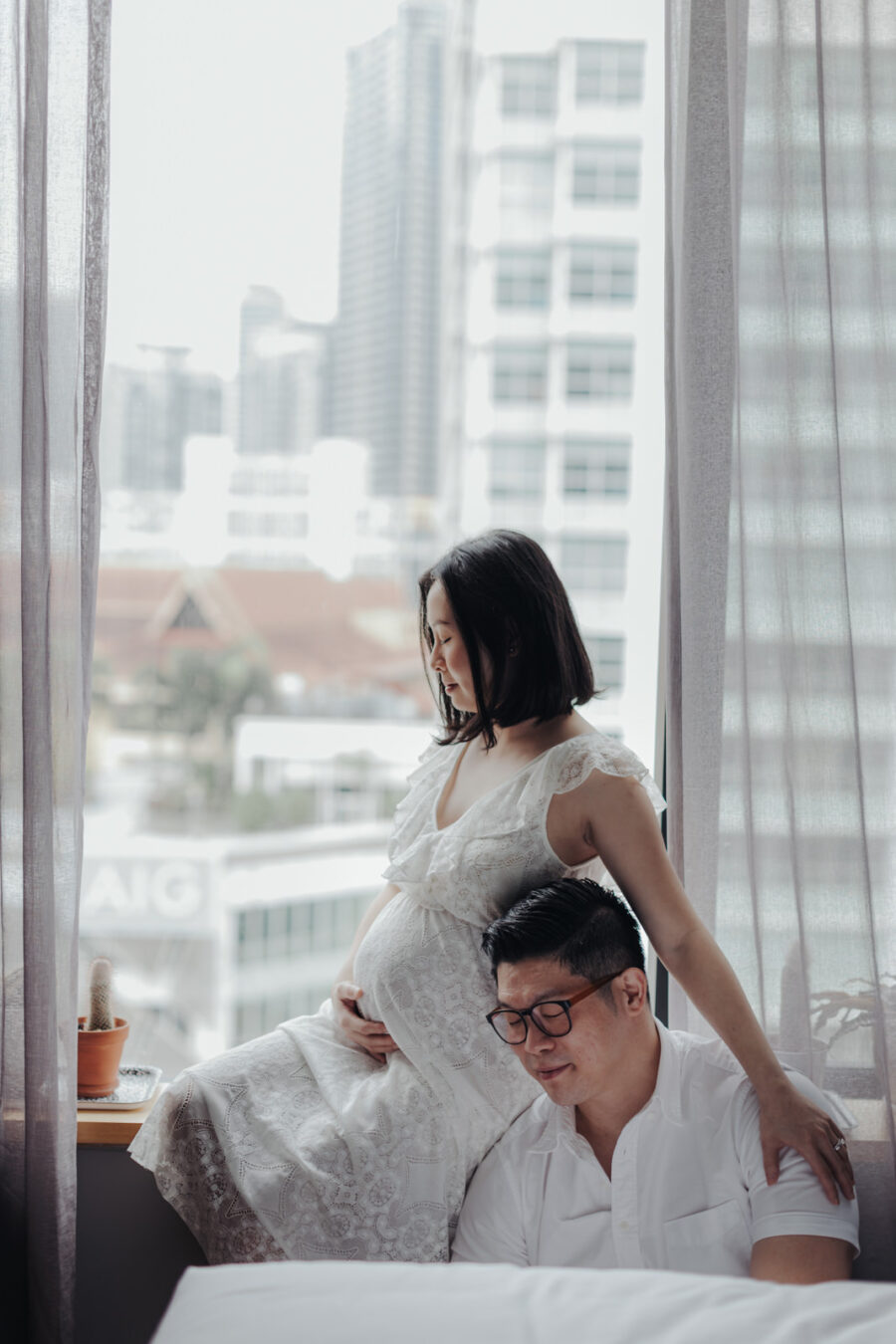 A pregnant wife sitting on a bed by the window in a white lace dress, gently cradling her belly and looking down with a peaceful smile. The cityscape is visible through the window, enhancing their maternity photo session.