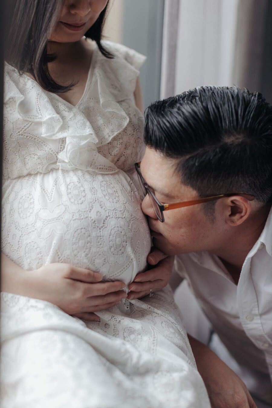 A close-up of a husband lovingly resting his head against his pregnant wife's belly, with both of them smiling gently. The wife is wearing a white lace dress, capturing an intimate moment in their maternity photo session.