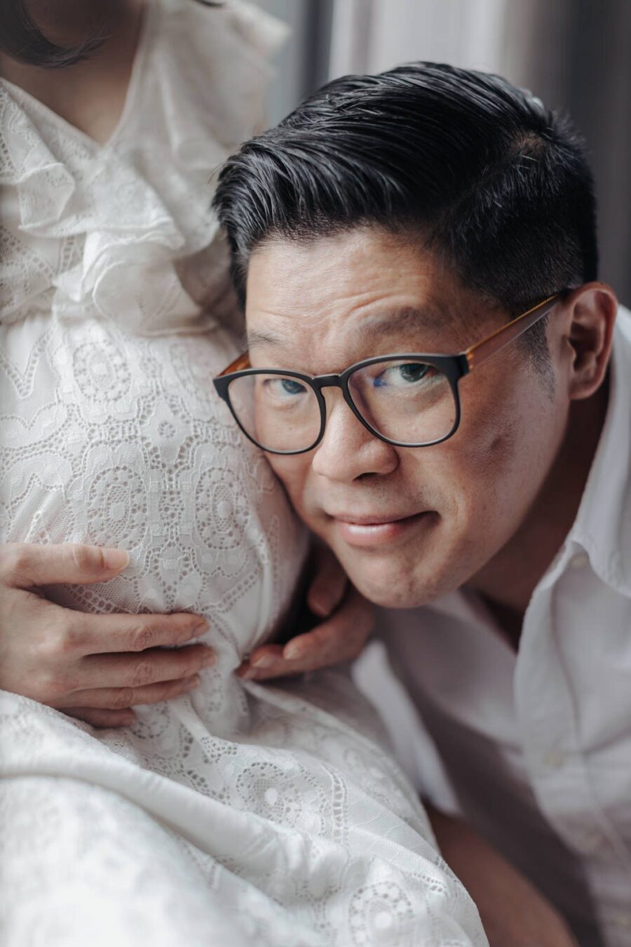 A close-up of a husband lovingly resting his head against his pregnant wife's belly, with both of them smiling gently. The wife is wearing a white lace dress, capturing an intimate moment in their maternity photo session.