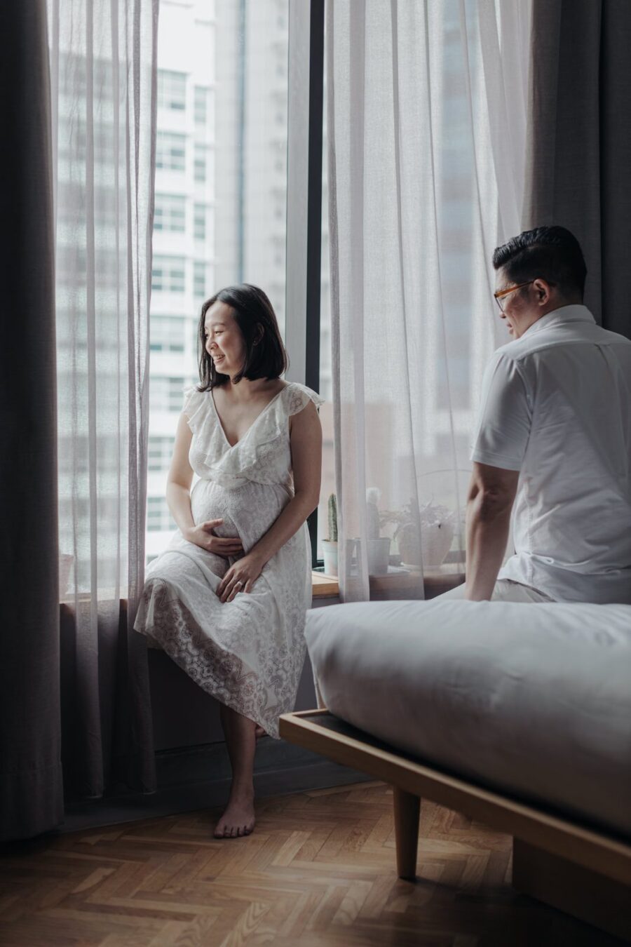 A pregnant wife in a white lace dress sitting on a windowsill, gently cradling her belly, with her husband sitting on the floor beside her. They both have a calm and reflective expression during their maternity photo session.