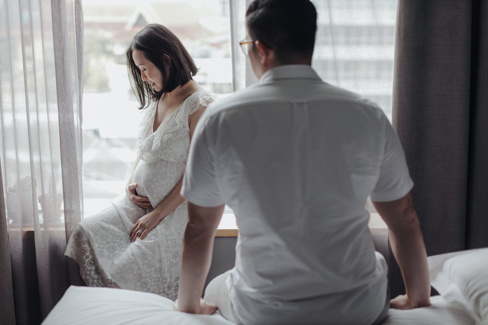 A pregnant woman in a white lace dress is sitting by the window, looking down and cradling her belly. Her husband is sitting on the bed behind her, watching her with a gentle smile.