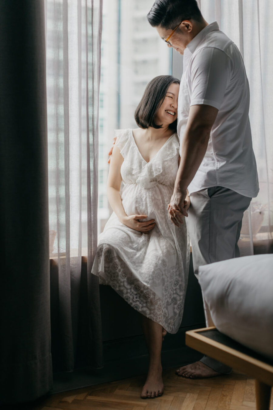 A happy pregnant woman in a white lace dress is sitting by the window, smiling at her husband who is standing beside her. They are holding hands and looking at each other lovingly.