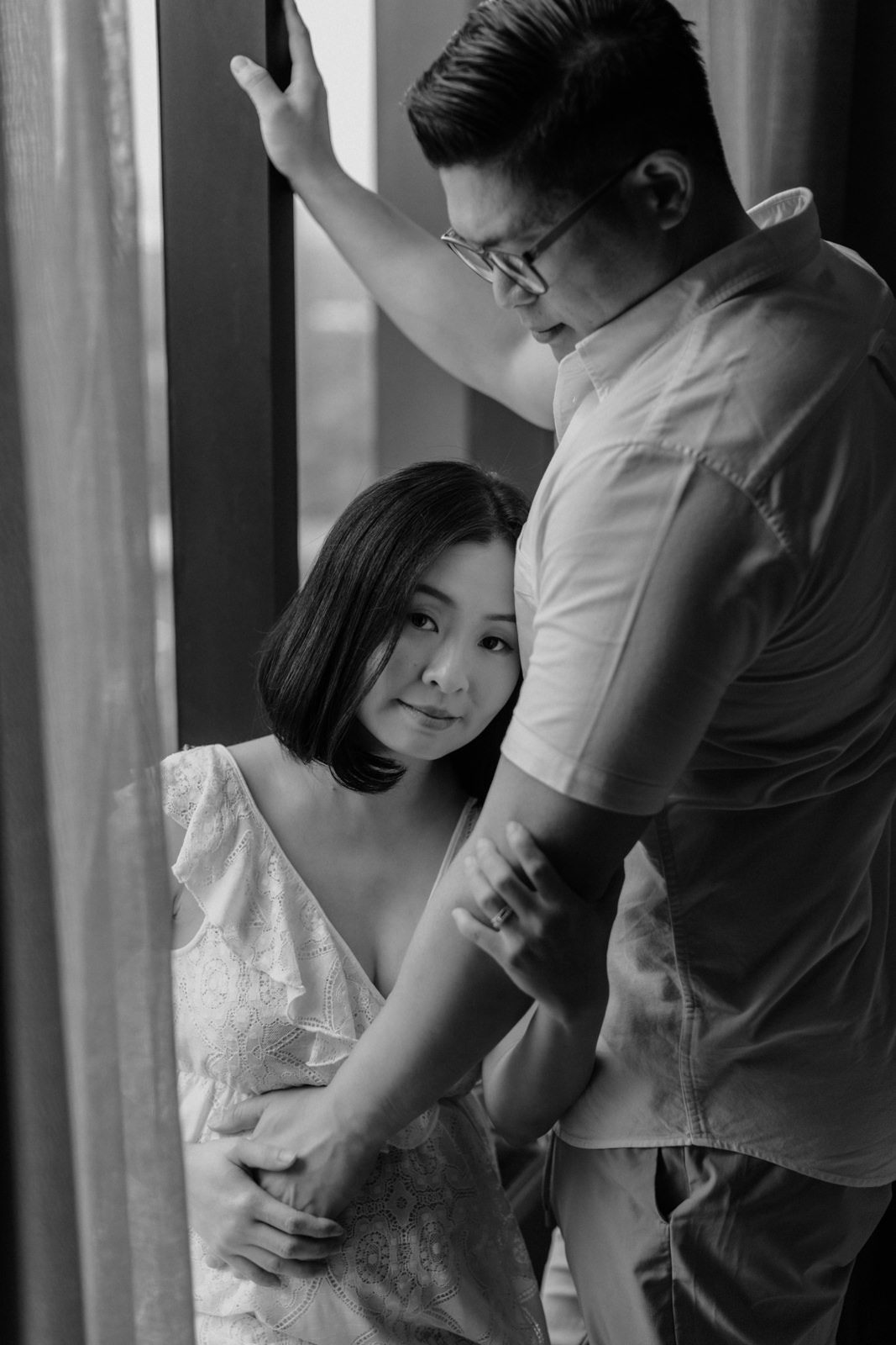 A black-and-white photo from a maternity photo session at KLoe Hotel, showing a tender moment between a husband and his pregnant wife. The wife is gently leaning her head against her husband's chest while he holds her and places a protective arm on the wall.