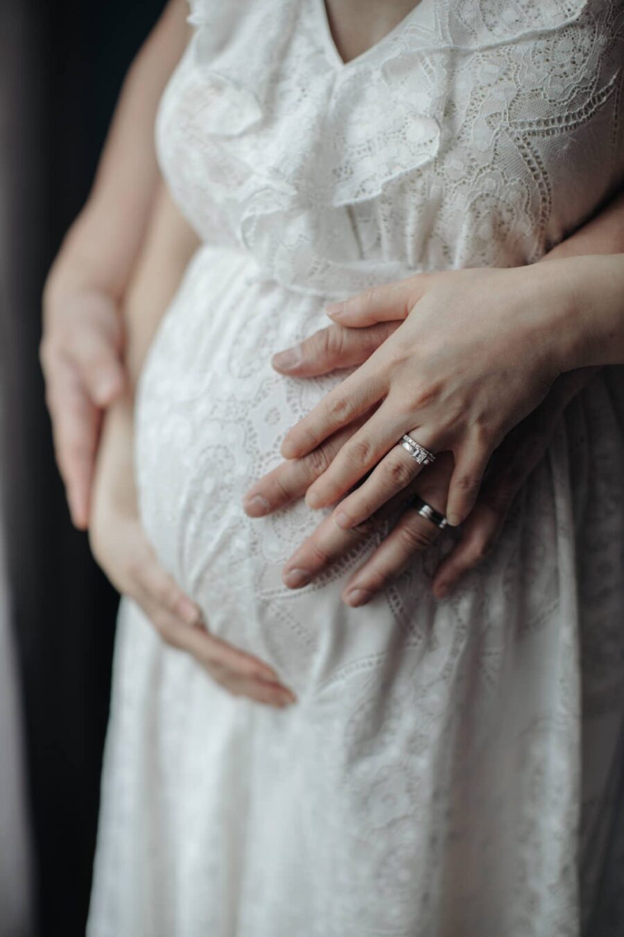 A close-up image of a pregnant woman's belly in a white lace dress. Hands of the woman and her husband are gently cradling the belly, showing their wedding rings.