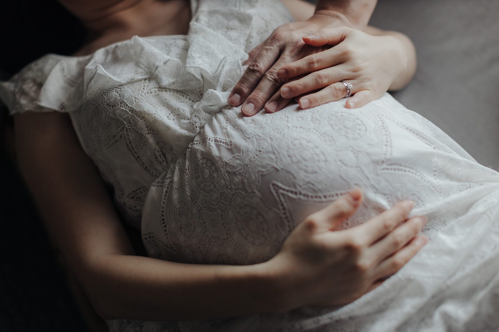 A pregnant woman lying down in a white lace dress. Her and her husband's hands are placed gently on her belly.