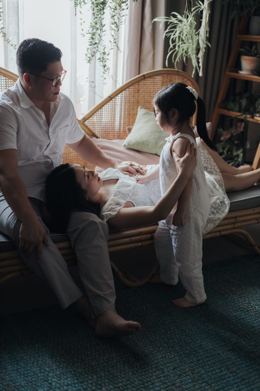 A husband sitting on a couch with his pregnant wife lying down, her head resting on his lap. Their young daughter stands beside them, holding her mother's hand, creating a tender family moment captured in their maternity photo session.