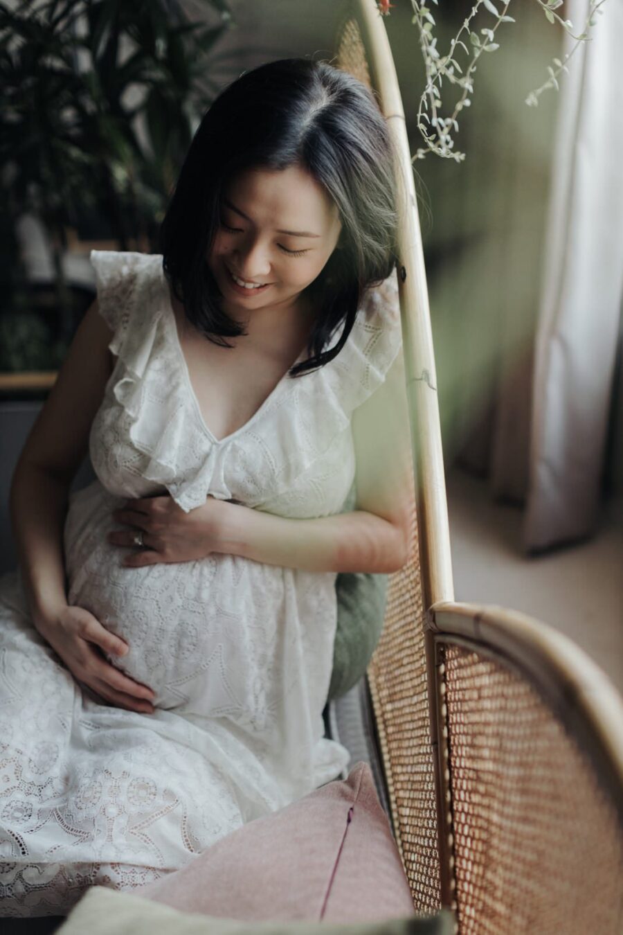 A pregnant wife in a white lace robe kneeling on a couch surrounded by greenery, gently holding her belly and looking down with a peaceful smile.