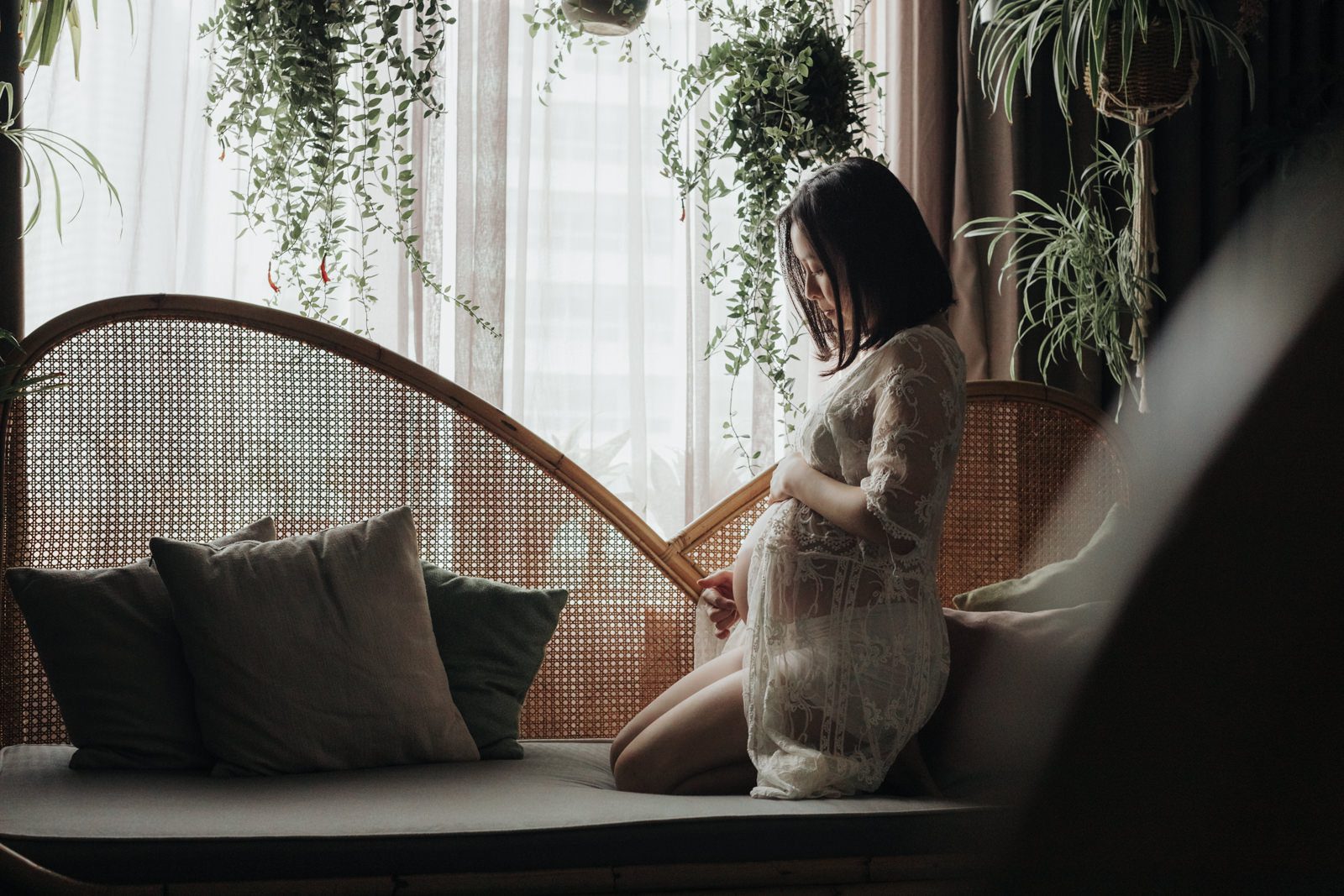 A pregnant wife in a white lace robe kneeling on a ratten couch surrounded by greenery, gently holding her belly and looking down with a peaceful smile. KLoe Hotel.