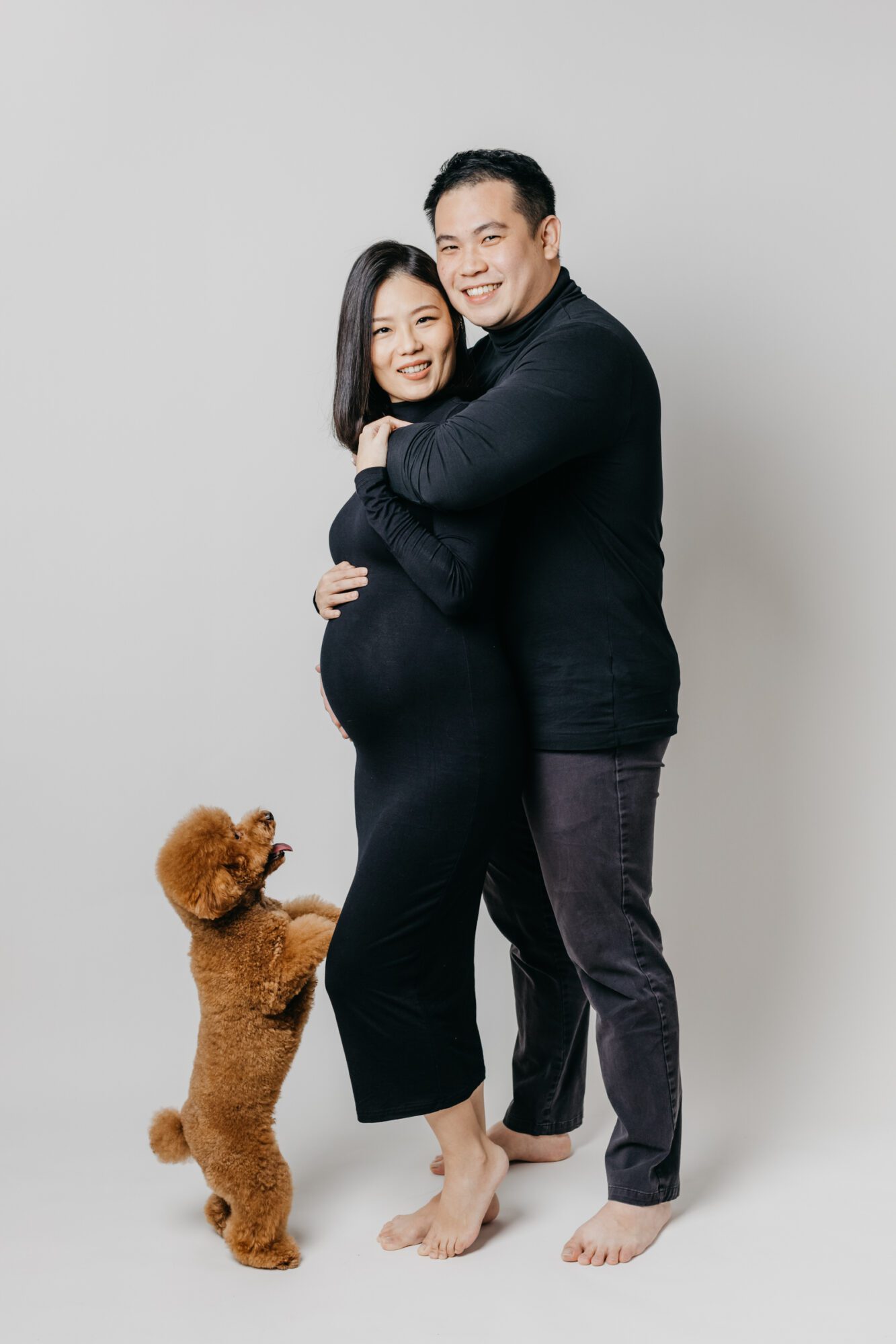 Mother and father hugging with their poodle, Milo, standing on hind legs during a maternity photo session