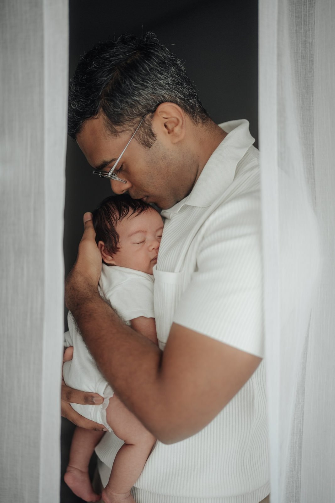 Father tenderly kissing baby Cleo’s head, capturing a heartfelt baby portrait in a warm portrait at home session.