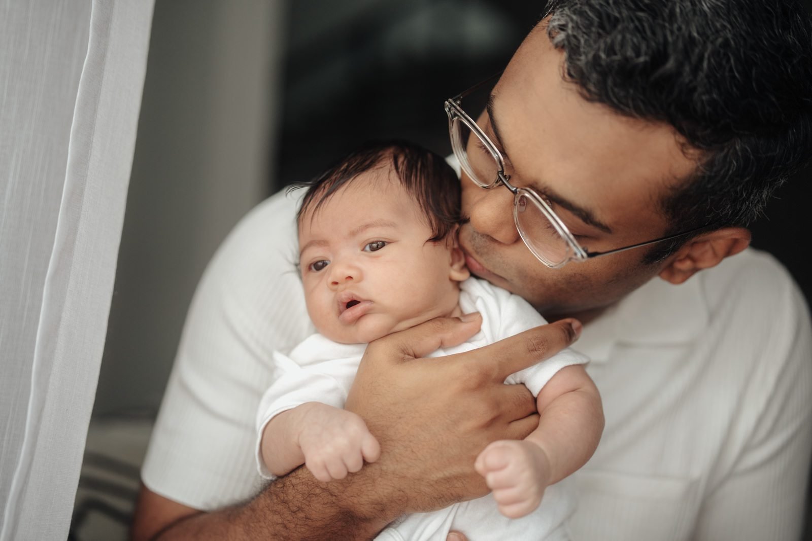 Father tenderly kissing baby Cleo’s head, capturing a heartfelt baby portrait in a warm portrait at home session. Cliff Choong Photography